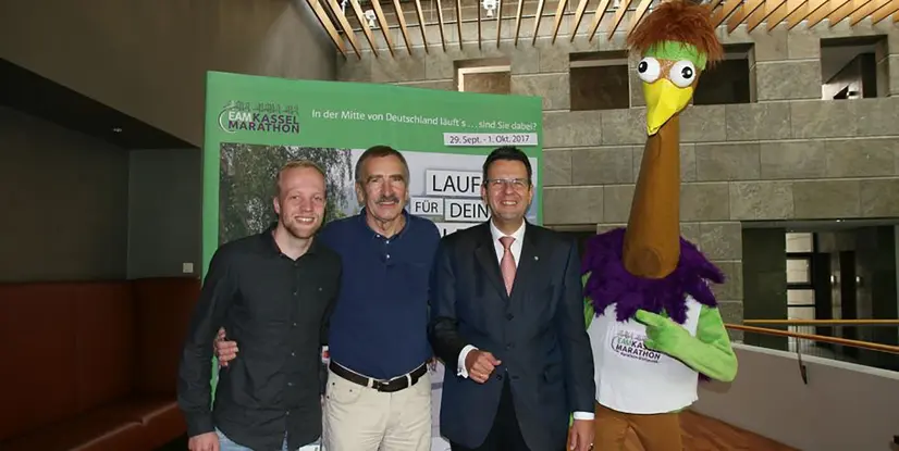 Vier Personen stehen zusammen, darunter ein Mensch in einem bunten Kostüm mit einem langen Hals und einem Gockelkopf, vor einem Marathon-Plakat.