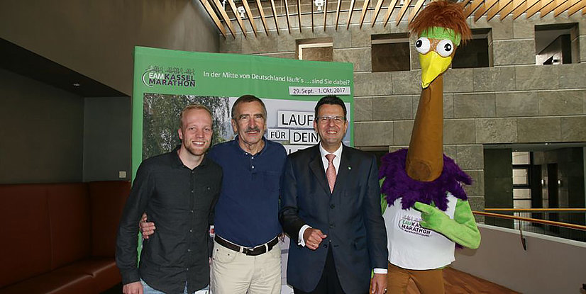 Vier Personen stehen zusammen, darunter ein Mensch in einem bunten Kostüm mit einem langen Hals und einem Gockelkopf, vor einem Marathon-Plakat.