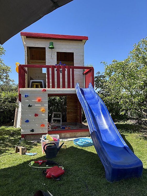 Buntes Spielhaus mit Rutsche und Kletterwand im Garten, umgeben von Spielzeug und Pflanzen.