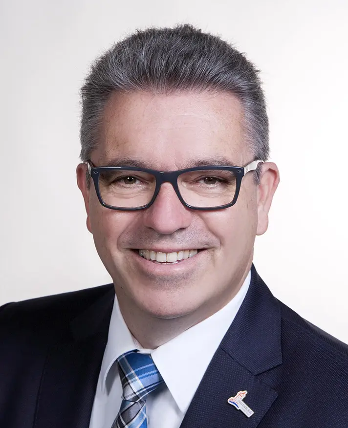 Michael Lotz, Bildrechte Fotostudio Bildschöne Momente Porträt eines lächelnden Mannes mit Brille, grau-gelocktem Haar, Anzug und Krawatte, vor einem neutralen Hintergrund.