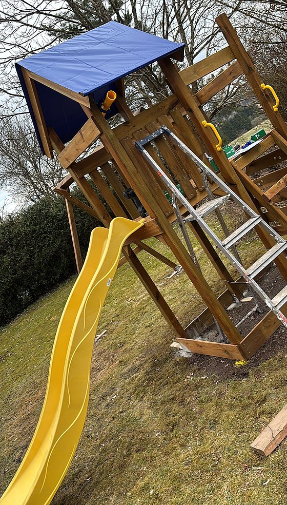 Spielplatzgerät mit Holzstruktur, blauer Decke, Geländerrutsche und Kletterleiter im Garten.