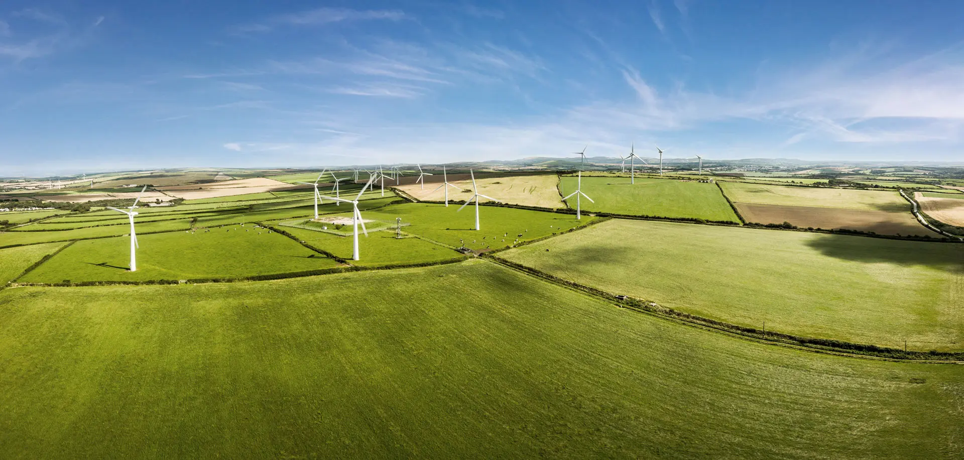 Luftaufnahme von Windrädern, die bei Sonnenschein zwischen Wiesen und Feldern stehen.