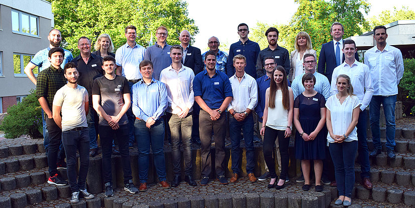 Gruppenfoto von 21 Personen im Freien, die auf einer Steinterrasse stehen, umgeben von Bäumen und Gebäuden.