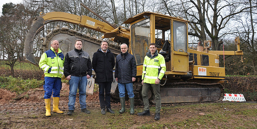 Fünf Personen stehen vor einem gelben Baumaschinenfahrzeug in einer ländlichen Umgebung.