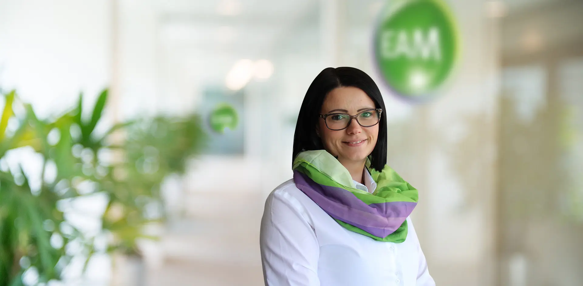 Porträt einer Frau mit Brille und buntem Schal, lächelnd in einem modernen Büro mit Pflanzen im Hintergrund.