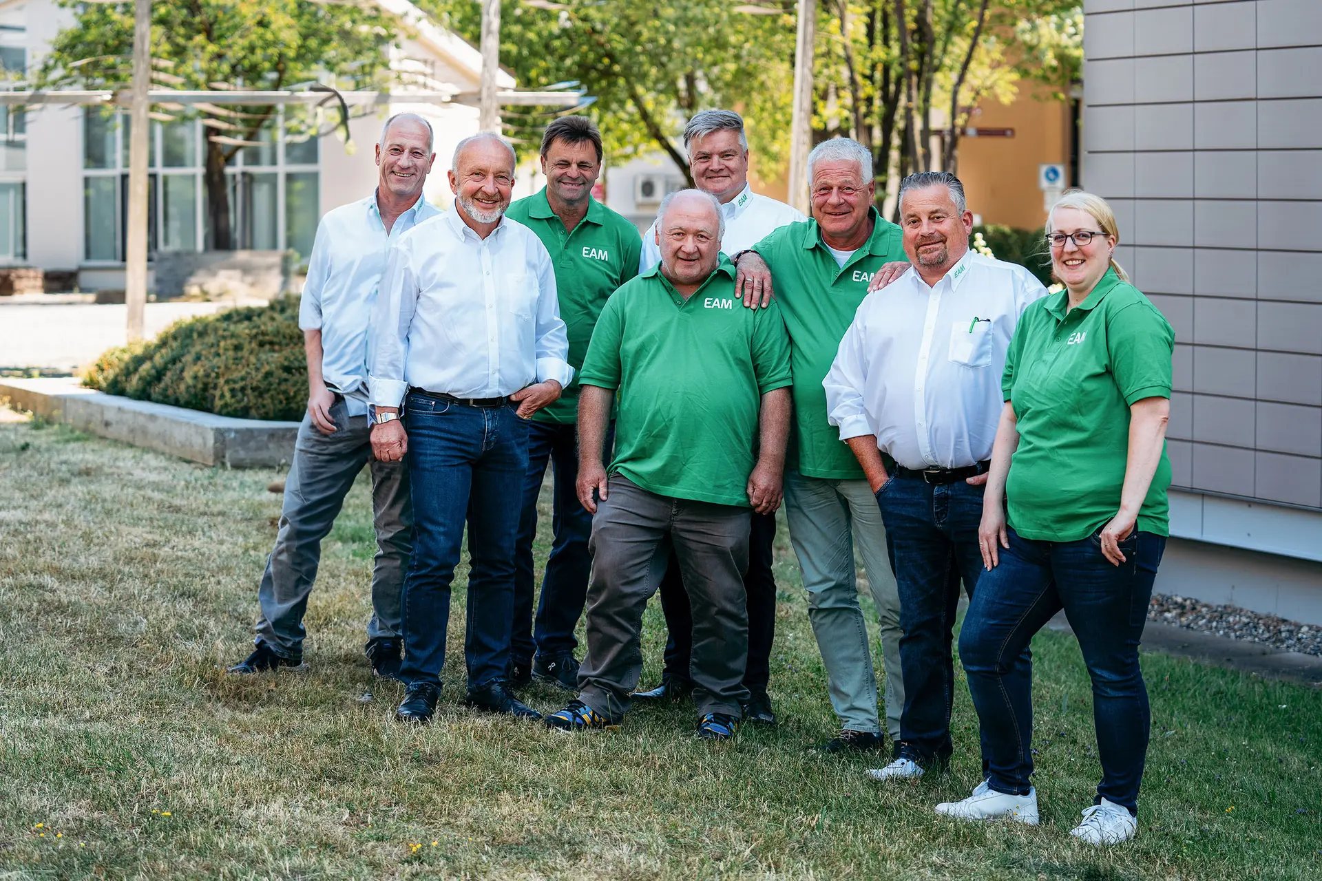 Eine Gruppe von neun Personen, einige in grünen EAM-Poloshirts, posiert lächelnd für ein Gruppenfoto im Freien.