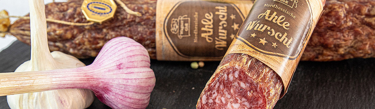 Ein Brett mit einer hübsch angerichteten Ahle Wurst, umgeben von Knoblauch, Tomaten und Gewürzen.