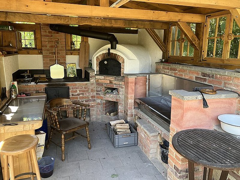 Barrierefreie Außenküche mit Holzofen, Tisch, Stühlen und verschiedenen Kochutensilien, umgeben von Natur.