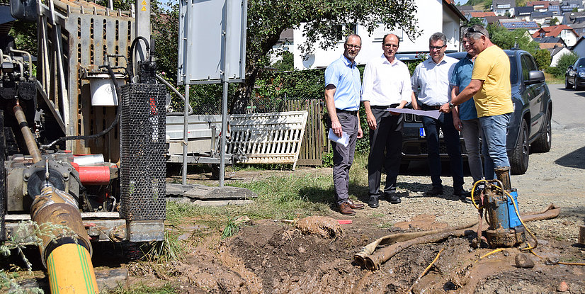 Vier Personen besprechen eine Baustelle neben einem Bagger in einem Wohngebiet, umgeben von Erde und Rohren.