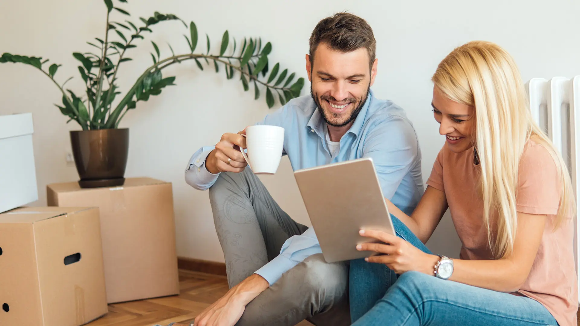Ein Mann und eine Frau sitzen auf dem Boden, trinken Kaffee und schauen gemeinsam auf ein Tablet. Umgeben von Umzugskartons.