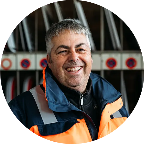 Lächelnder Mann in orangefarbener Arbeitsjacke vor einem Regal mit Schildern.