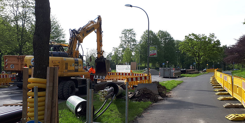 Bauarbeiten an einer Straße mit Bagger, Absperrgittern und Bauleitern in orangefarbenen Warnwesten.