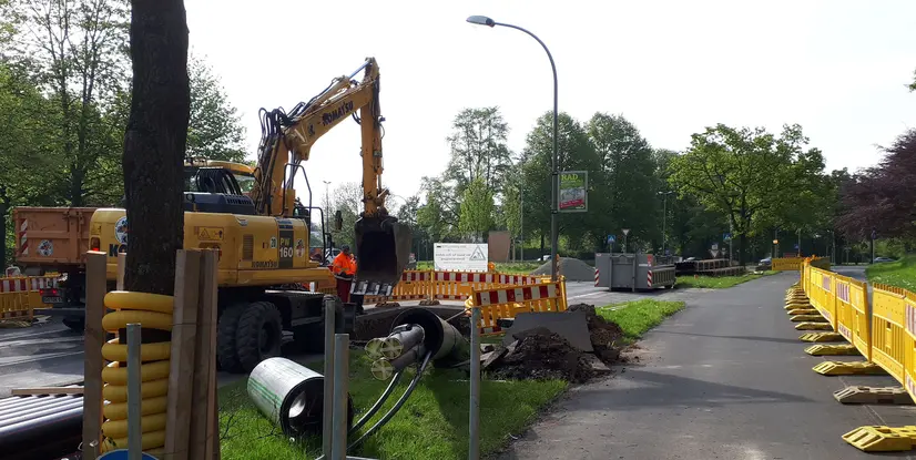 Bauarbeiten an einer Straße mit Bagger, Absperrgittern und Bauleitern in orangefarbenen Warnwesten.