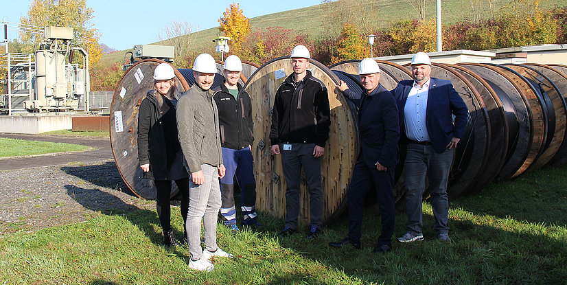 Sechs Personen mit Helm stehen neben großen Kabeltrommeln auf einer Wiese, im Hintergrund sind Arbeitseinrichtungen sichtbar.