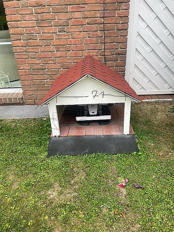 Ein kleiner, weißer Hundehütte mit rotem Dach steht auf einem Rasen vor einer Backsteinwand.