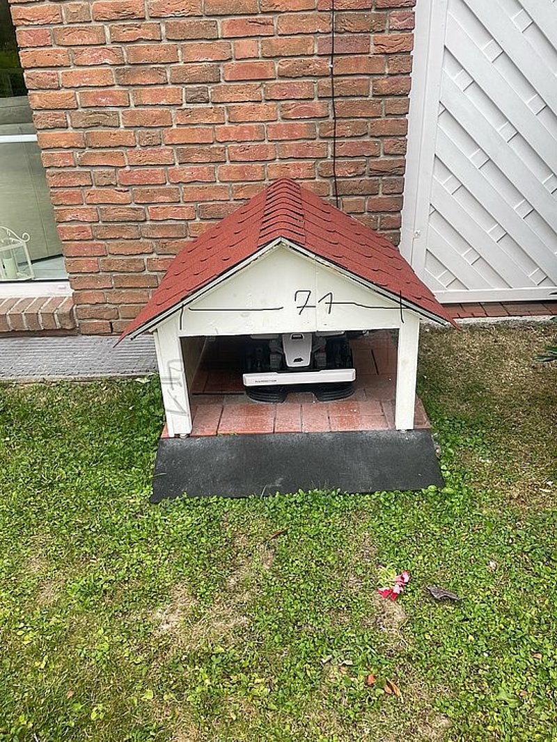 Ein kleiner, weißer Hundehütte mit rotem Dach steht auf einem Rasen vor einer Backsteinwand.