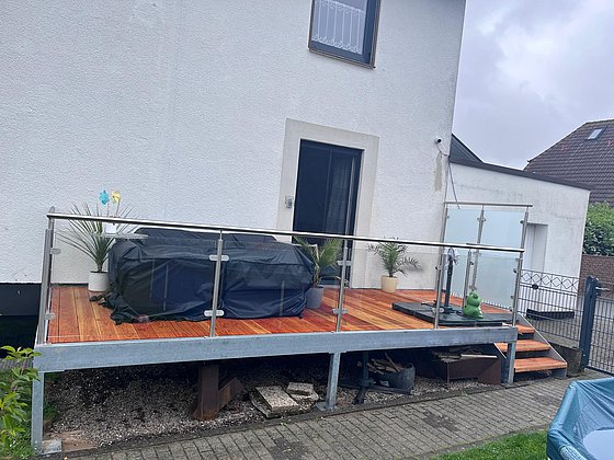 Terrasse mit Holzbelag, Pflanzen, abgedecktem Whirlpool und Geländer, angrenzend an ein weißes Wohnhaus.