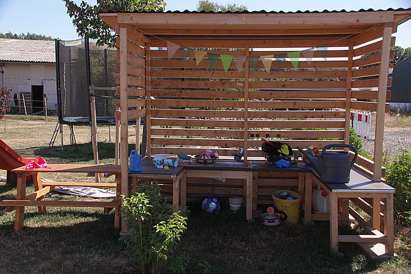 Holzschutzunterstand mit Arbeitsfläche, Spielzeug und Gießkanne, umgeben von Rasen und Bäumen.