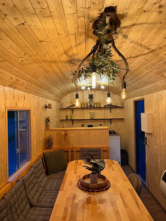 Gemütlicher Innenraum eines Holzhauses mit Holztisch, bequemen Sitzgelegenheiten und dekorativer Beleuchtung.