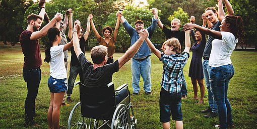 Eine Gruppe von Menschen unterschiedlichen Alters und mit unterschiedlichen Hautfarben steht im Freien auf einer Wiese in einem Kreis und hält sich an den Händen; eine Person sitzt im Rollstuhl. Im Hintergrund sind Bäume und Grünflächen bei Tageslicht zu sehen.