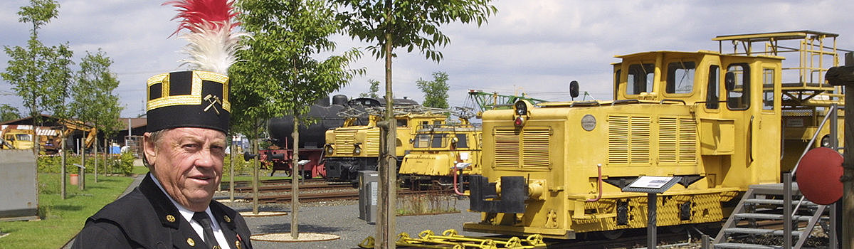 Eine Person in schwarzem Anzug mit Kopfbedeckung steht in einem Freilichtmuseum für Eisenbahnen mit gelben Lokomotiven im Hintergrund.