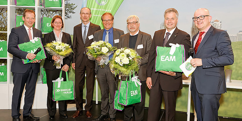 Gruppenfoto von sieben Personen bei einer Veranstaltung, alle halten Blumensträuße und Taschen mit EAM-Logo.