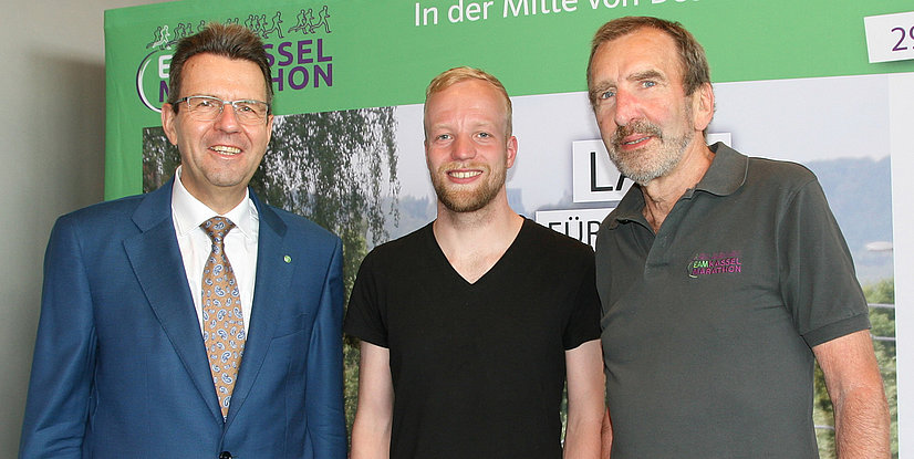 Drei Männer stehen lächelnd vor einem Plakat zum EAM Kassel Marathon, umgeben von grüner Szenerie.