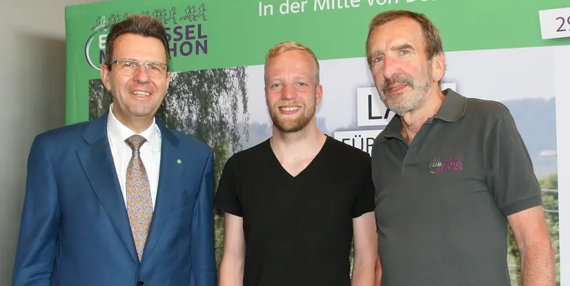 Drei Männer stehen lächelnd vor einem Plakat zum EAM Kassel Marathon, umgeben von grüner Szenerie.