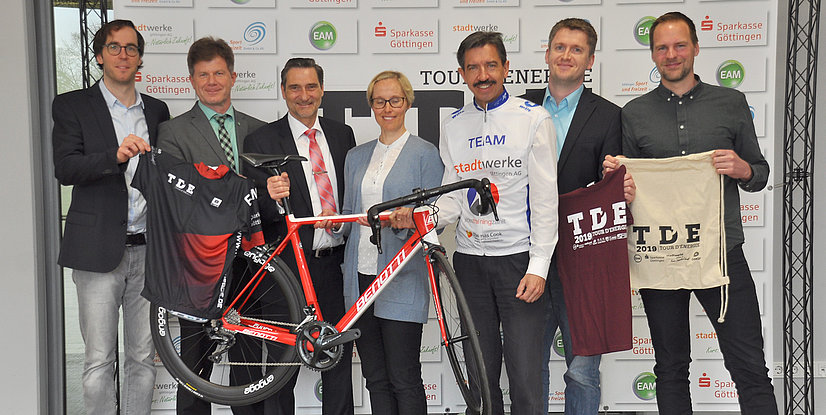 Gruppenbild von sieben Personen bei der Präsentation der Tour d'Energie mit Rad und T-Shirts im Hintergrund.