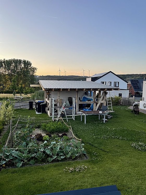 Holzschuppen im Garten mit Möbeln, Mülltonnen und Pflanzen, umgeben von einer Wiese bei Sonnenuntergang.