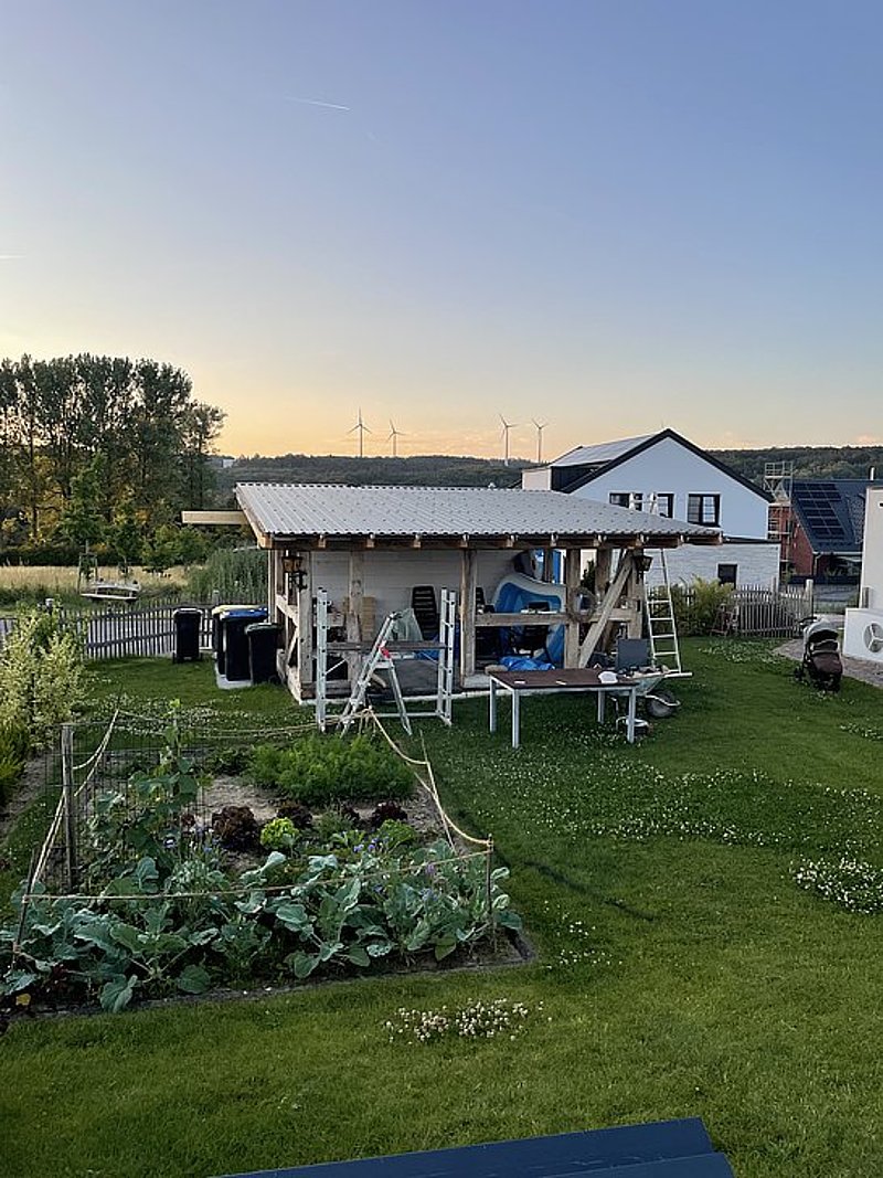 Holzschuppen im Garten mit Möbeln, Mülltonnen und Pflanzen, umgeben von einer Wiese bei Sonnenuntergang.