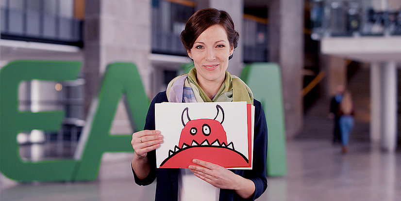 Eine Frau mit einem bunten Schal hält ein Blatt Papier mit einer roten Monsterzeichnung in die Kamera. Im Hintergrund ist ein grünes Logo zu sehen.