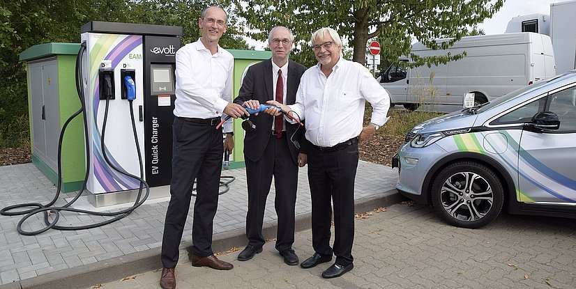 Drei Männer an einer Ladestation für Elektrofahrzeuge halten ein Kabel und lächeln in die Kamera.