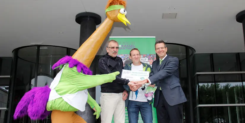 Drei Männer und ein Maskottchen überreichen eine Urkunde beim Kassel Marathon, vor einem modernen Gebäude.