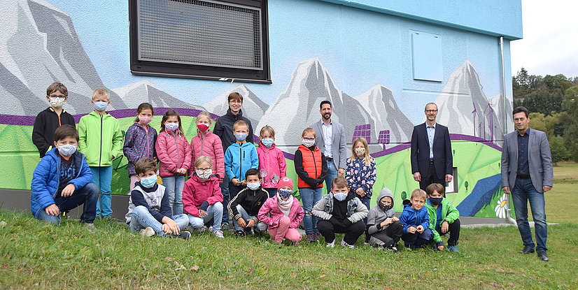 Gruppe von Kindern und Erwachsenen vor einem Gebäude mit einem Graffiti das eine Berglandschaft zeigt.