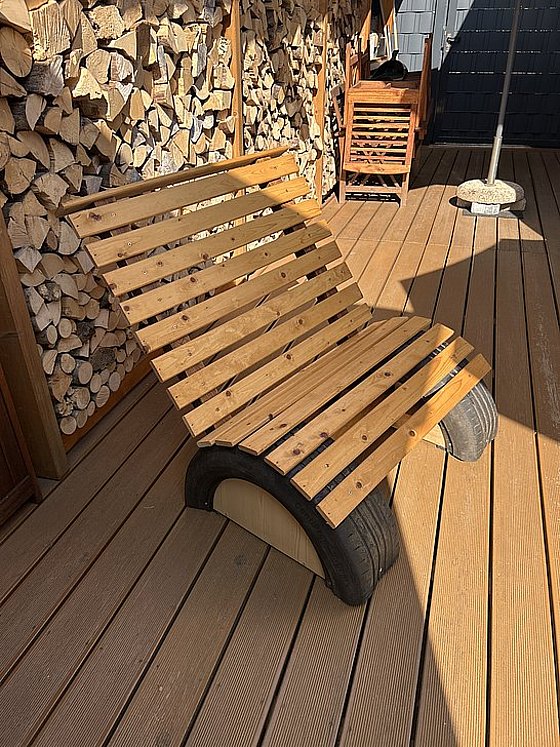 Ein Holzliegestuhl mit Reifenfüßen steht auf einer Holzterrasse, im Hintergrund Holzstapel und eine Sonnenschirmbasis.