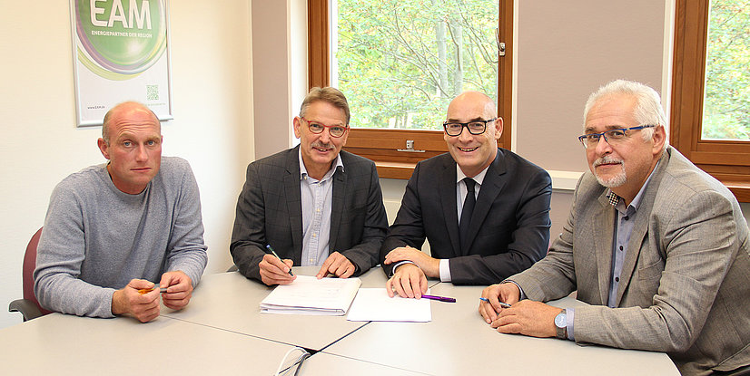 Vier Personen sitzen an einem Tisch, arbeiten an Dokumenten und besprechen etwas. Im Hintergrund ist ein Fenster sichtbar.