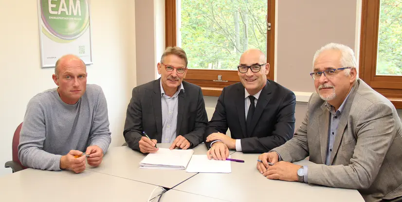 Vier Personen sitzen an einem Tisch, arbeiten an Dokumenten und besprechen etwas. Im Hintergrund ist ein Fenster sichtbar.