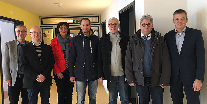 Gruppe von sieben Personen in einem Flur, lächelnd, in formeller Kleidung. Hintergrund mit gelben und weißen Wänden.