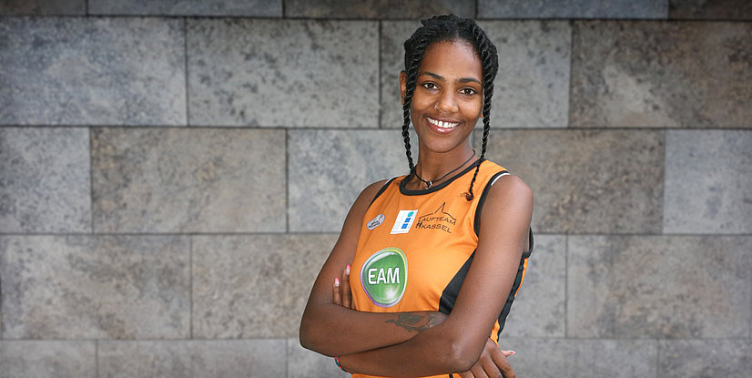 Die Marathonläuferin Melat Kejata, in orangefarbenem Trikots mit EAM-Logo mit Armen verschränkt vor einer grauen Mauer, bereit für den Wettkampf.