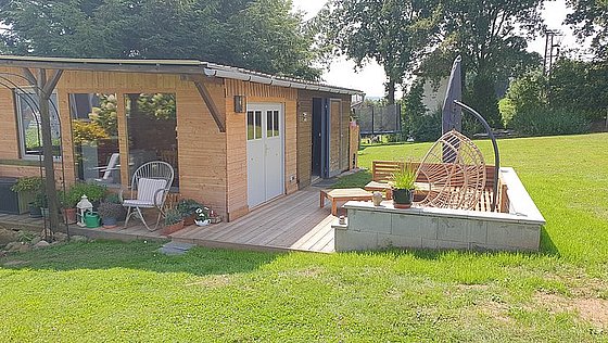 Holzhütte mit Terrasse, Gartenmöbeln und Rasen, umgeben von Bäumen und Pflanzen.
