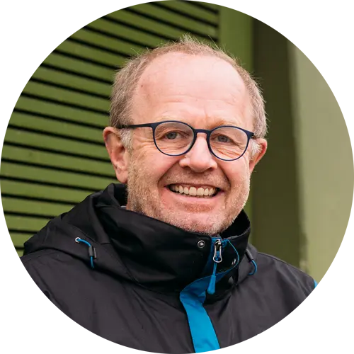 Mann mit Brille und lächelndem Gesicht, trägt eine schwarze Jacke mit blauen Akzenten, vor einer grünen Wand.