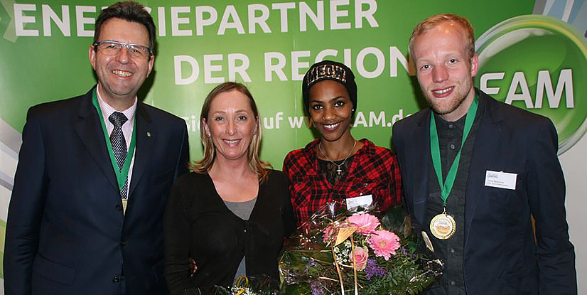 Vier Personen lächeln in einem festlichen Rahmen, eine hält Blumen, mit einem grünen Hintergrund und Medaillen um den Hals.