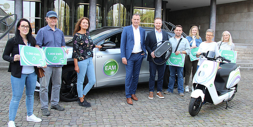 Gruppe von Menschen mit Schildern über 1.000 kWh vor einem E-Auto und einem Elektroroller, feiern nachhaltige Mobilität.