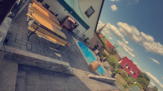 Ein Gartenbereich mit einem Pool, Holzbänken und Pavillon unter blauem Himmel mit Wolken.