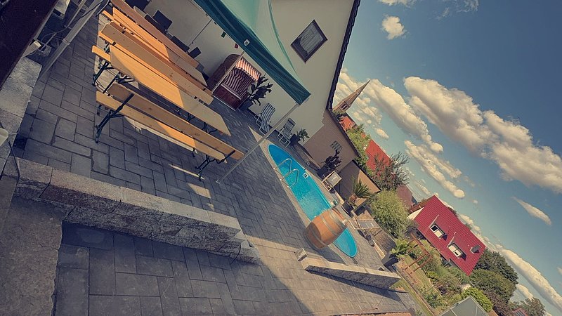 Ein Gartenbereich mit einem Pool, Holzbänken und Pavillon unter blauem Himmel mit Wolken.