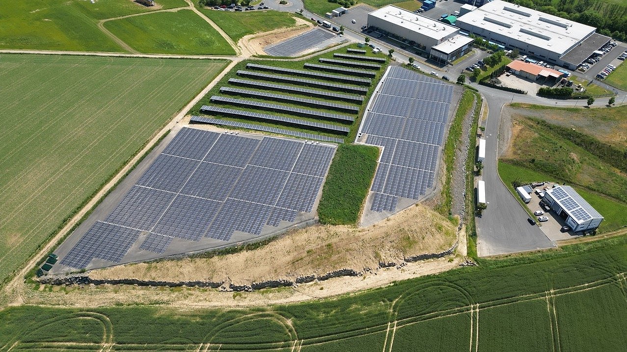  Luftaufnahme einer großen Solarpark-Anlage mit vielen Solarmodulreihen, umgeben von grünen Feldern und einem Industriegebiet.