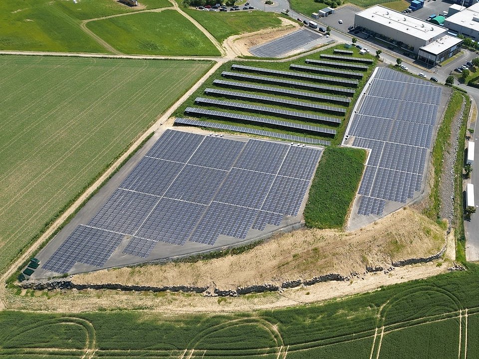 Luftaufnahme einer großen Solarpark-Anlage mit vielen Solarmodulreihen, umgeben von grünen Feldern und einem Industriegebiet.