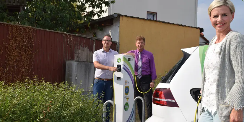 Drei Personen stehen neben einer Ladestation für Elektrofahrzeuge, ein Fahrzeug wird gerade aufgeladen.