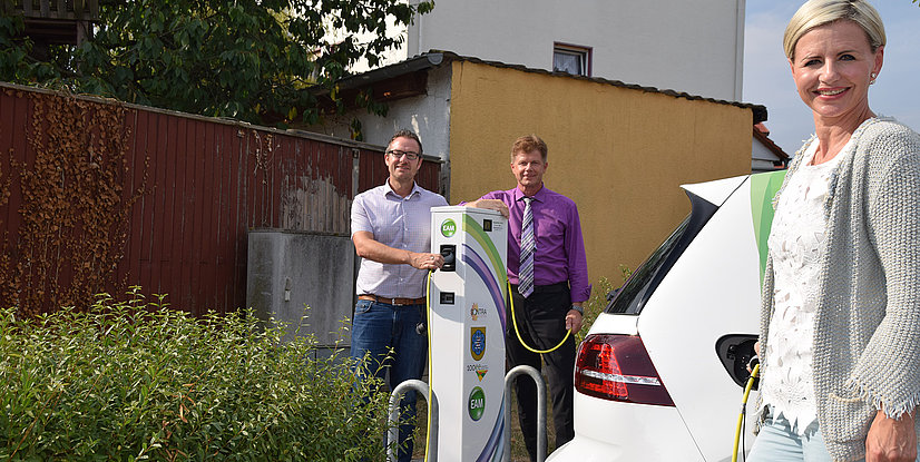 Drei Personen stehen neben einer Ladestation für Elektrofahrzeuge, ein Fahrzeug wird gerade aufgeladen.